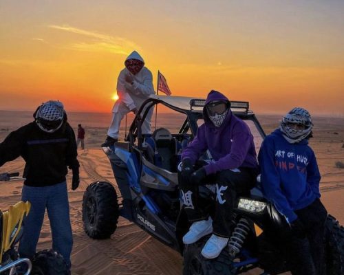 One dune desert safari buddy parked in the desert under an open sky, with four men sitting and standing on and around the buggy